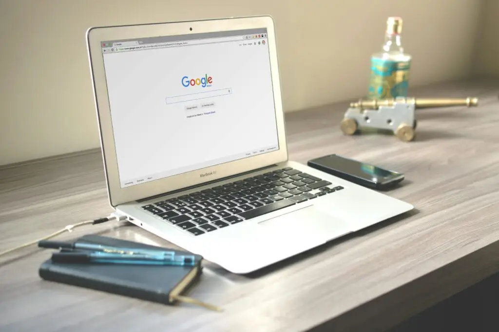A laptop displaying the Google homepage sits on a wooden desk. Nearby, a smartphone, a notebook, and two pens are arranged next to the laptop, alongside a decorative bottle and a small brass cannon. Elevate Web website designer toronto