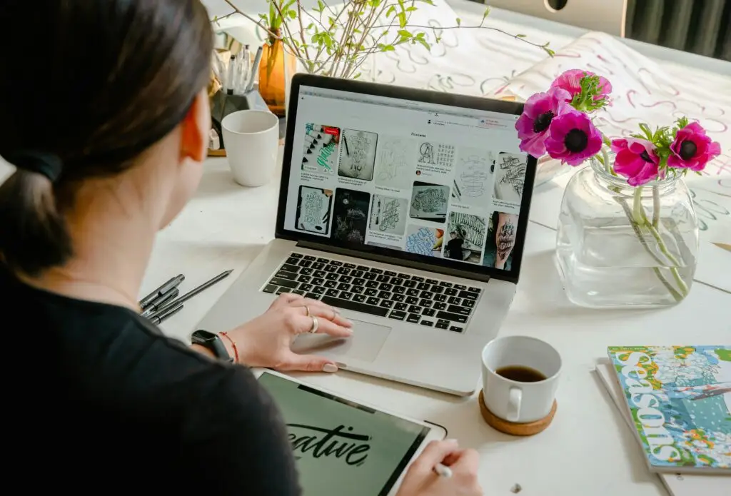 A person viewing a design website on a laptop at a table, with a tablet and coffee cup nearby. A vase of flowers is also on the table, alongside open magazines. The scene emphasizes small business website design. Elevate Web website designer toronto
