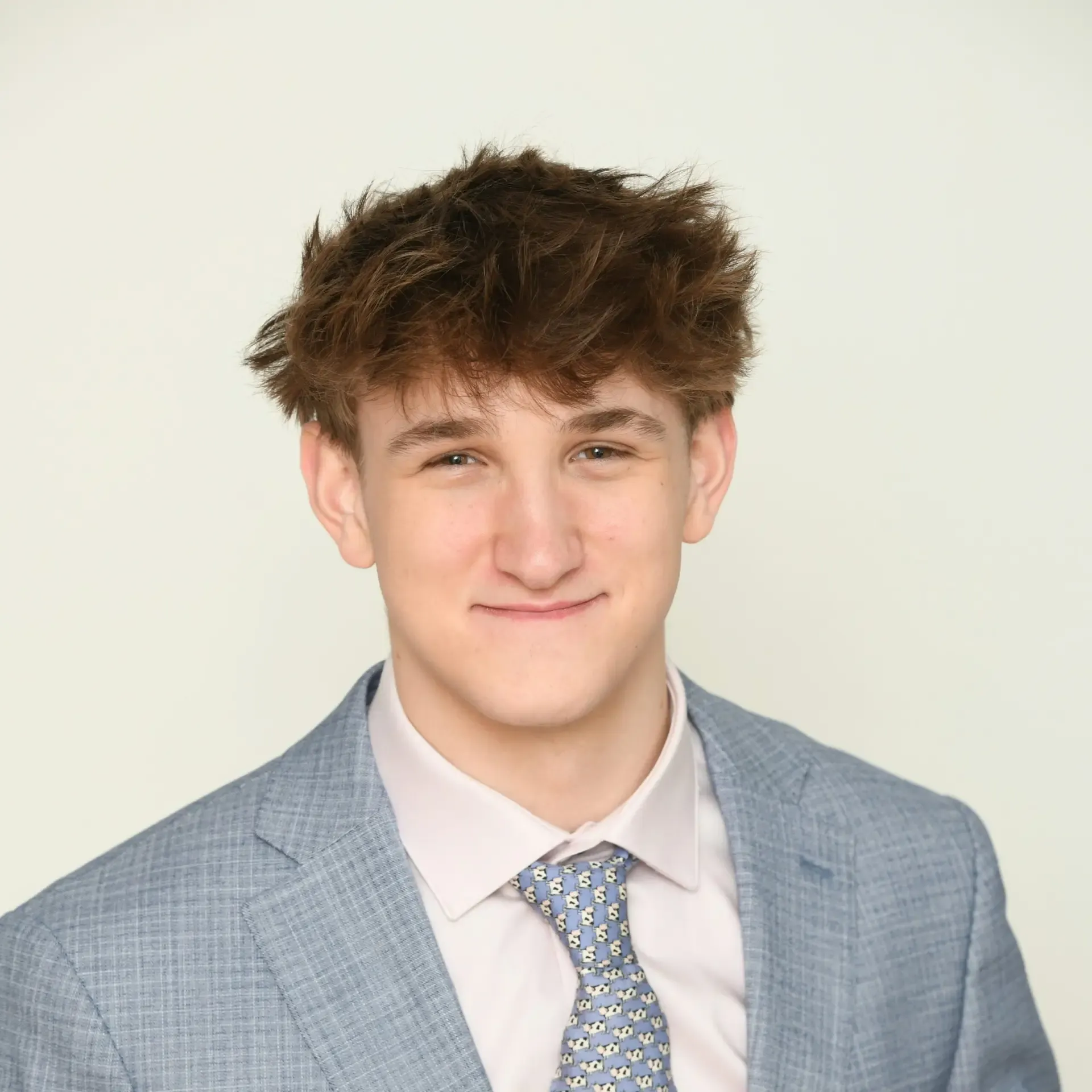 Young man in gray suit jacket and patterned tie smiling at camera with tousled brown hair against white background